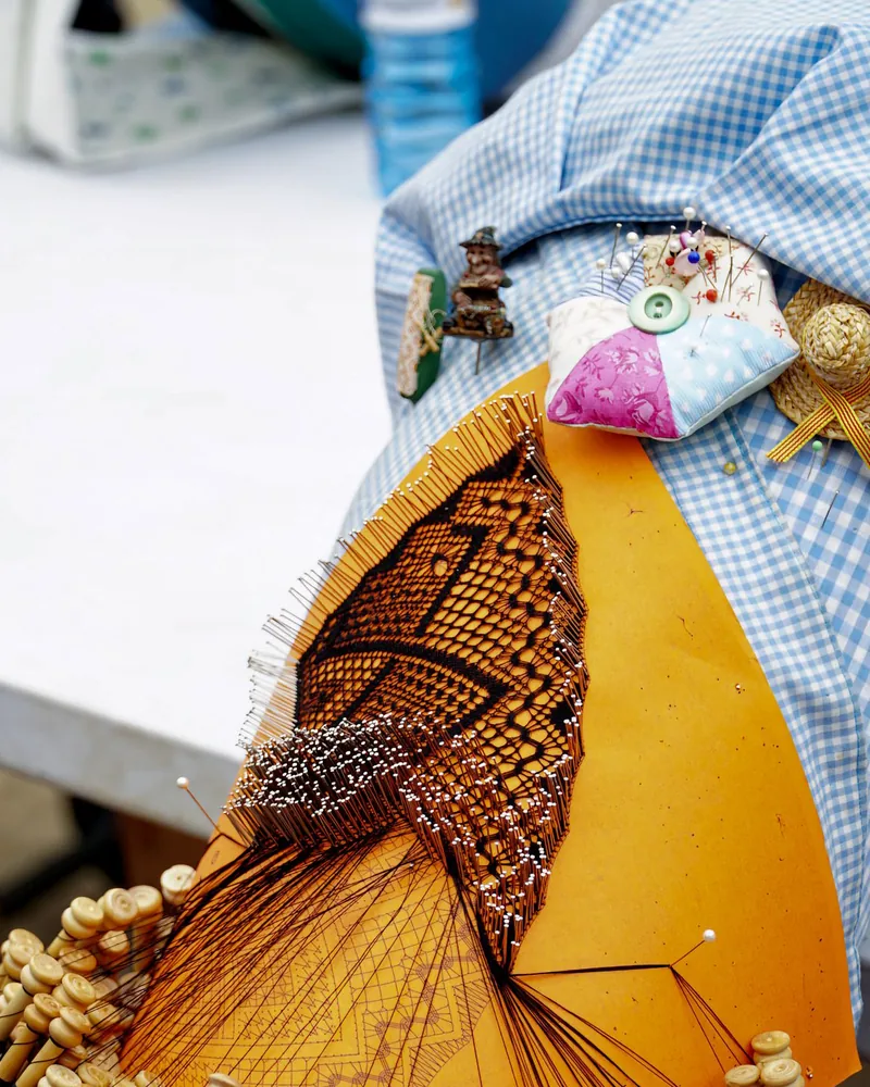 Encaje de bolillos a medio hacer sobre el picado del mundillo, con hileras de alfileres marcando el contorno del dibujo y los bolillos colgando