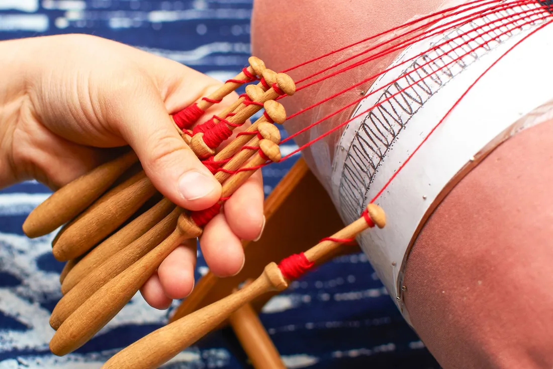 Manos sosteniendo varios bolillos de madera con hilo rojo sobre un mundillo cubierto por el picado y sujeto con alfileres